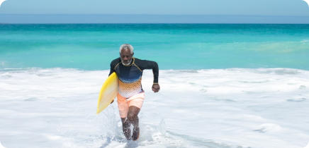 Photo of a senior man with a surfboard under his arm running through the shallow surf of a tropical beach