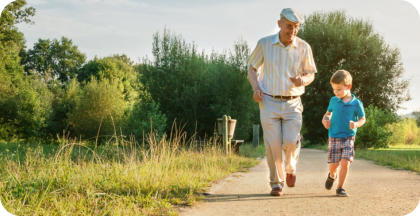 A senior man is jogging down an outdoor path with his school-age grandson
